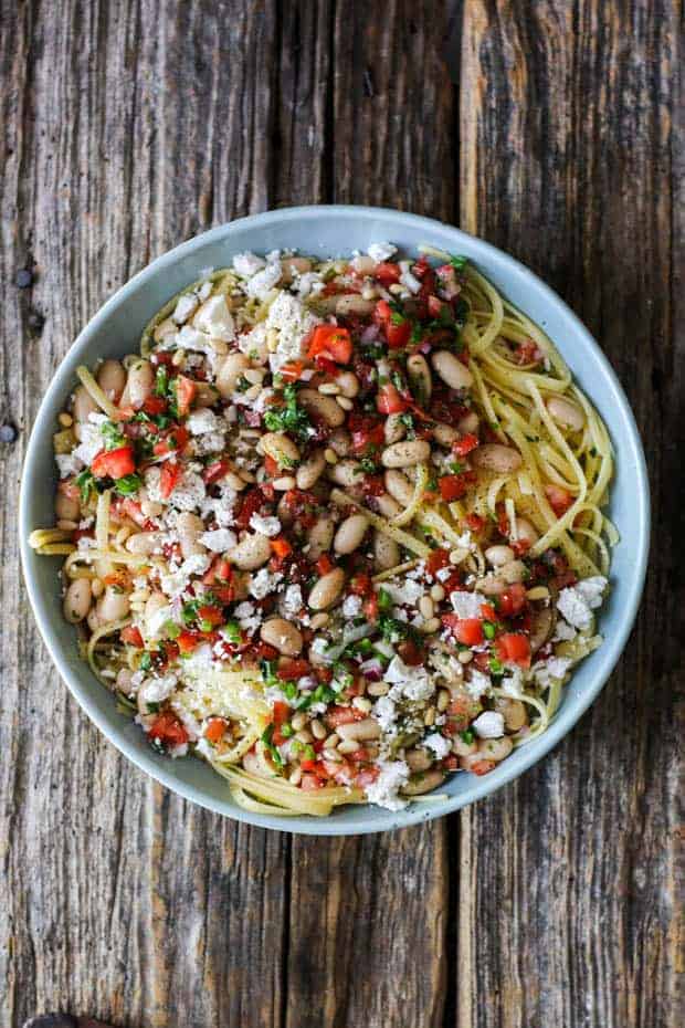A shallow blue bowl sits on a wooden table top. It has fettuccini noodles in it and they are topped with pico de gallo, white beans, and crumbled ricotta salata.