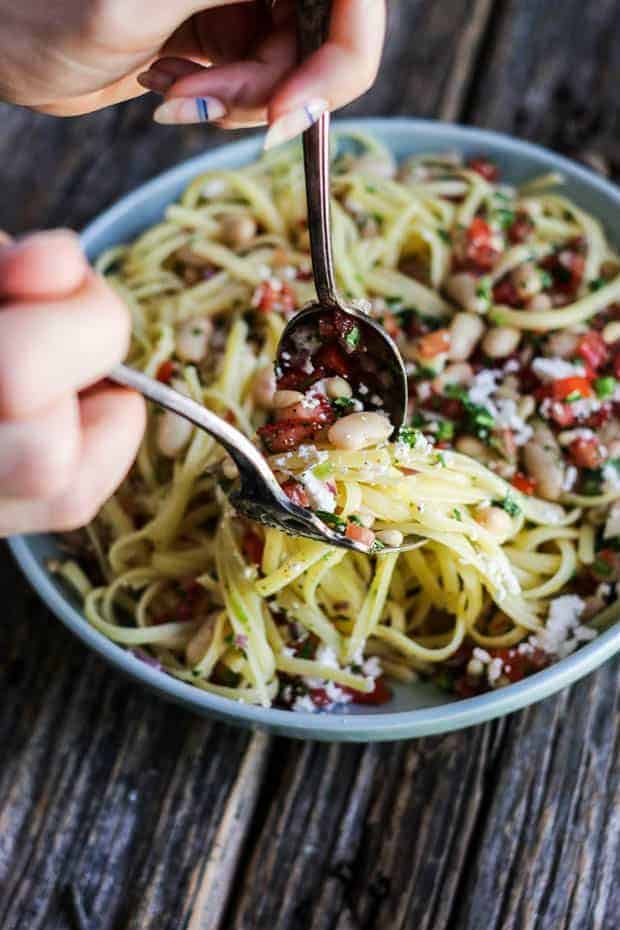 A shallow light blue bowl is on a wooden table, filled with fettuccini pasta, white beans, pico de gallo, and crumbled ricotta salata cheese.A shallow light blue bowl is on a wooden table, filled with fettuccini pasta, white beans, pico de gallo, and crumbled ricotta salata cheese. There is a woman's hands using a silver fork and spoon to pick pasta up out of the dish. 