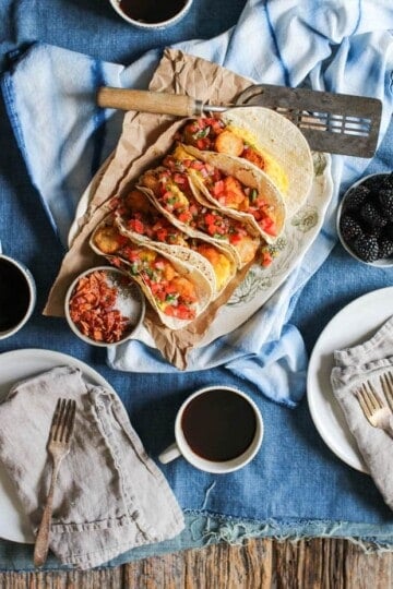 A platter full of breakfast tacos is on a table that is covered in a denim table cloth. There is a blue and white napkin on the table underneath the platter of tacos, mugs of coffee, and a bowl of blackberries on the table.