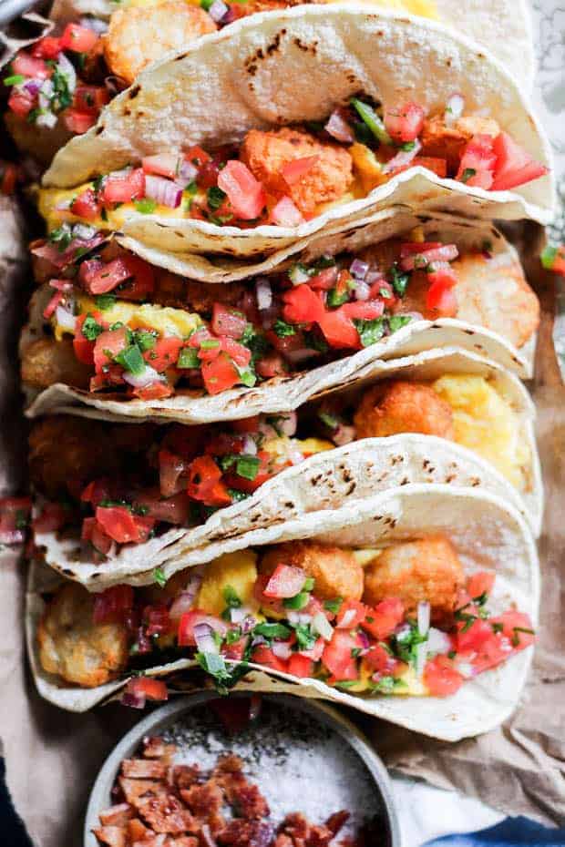 4 tacos, corn tortillas stuffed with scrambled eggs, tater tots, and pico de gallo. The tacos are arranged on a platter next to a bowl that has chopped bacon in it.