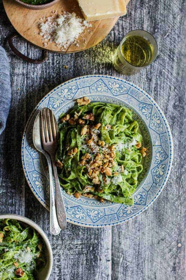 A large shallow bowl with a blue and cream decorative edge is piled high with noodles coated in a bright green pesto. There are walnuts and grated cheese on top and a fork and spoon on the side of the bowl. There is a glass if white wine next to the bowl.