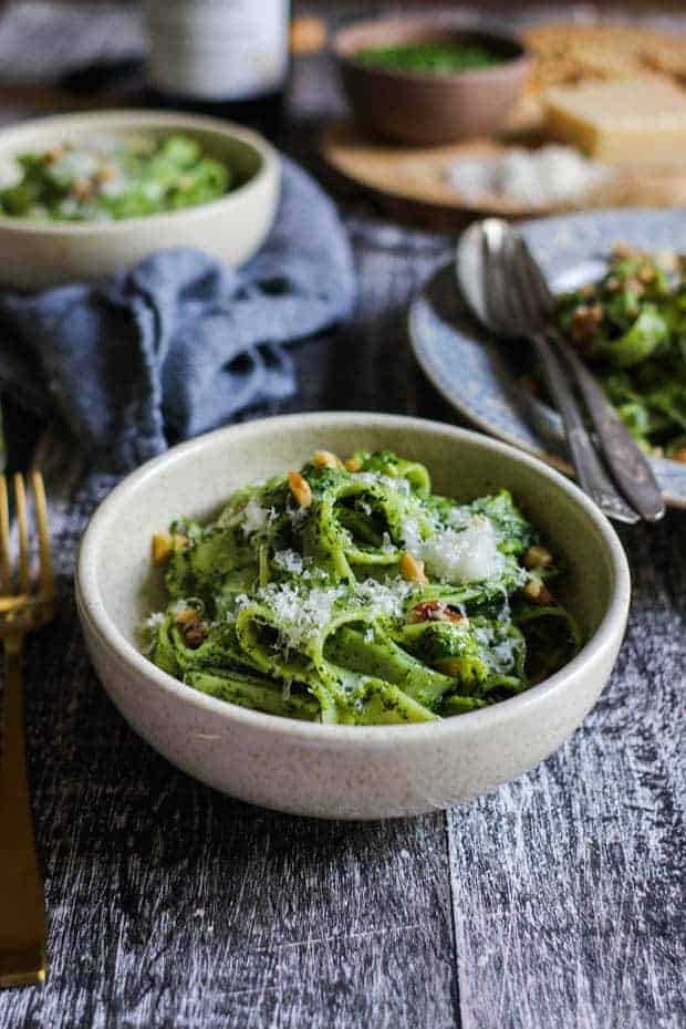 A cream colored bowl filled with vibrant green pesto dressed pasta. There are other bowls filled with pasta in teh background. They all have a sprinkle of chopped walnuts and grated cheese. 