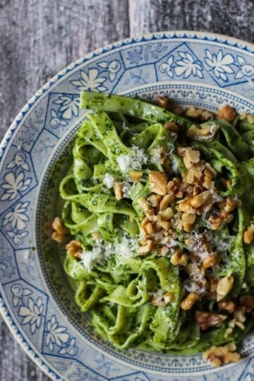 A close up image of a shallow blue and cream colored bowl filled with noodles coated in a bright green kale pesto. There are walnuts and grated cheese on top.
