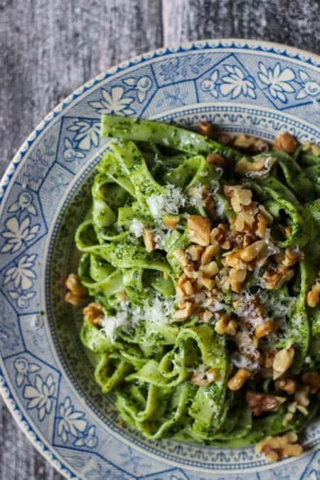 A close up image of a shallow blue and cream colored bowl filled with noodles coated in a bright green kale pesto. There are walnuts and grated cheese on top.