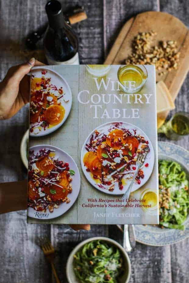 Janet Fletcher's new cookbook The Wine Country Table is being held over a table setting that is styled with walnut kale pesto pasta.