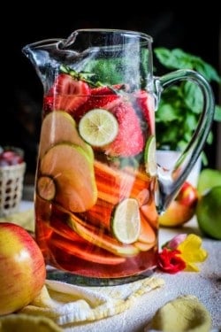 A glass pitcher filled with apple and berry rosé sangria and sliced red and green apples, halved berries, and lime wheels. There are fresh green and pink apples scattered on the table as well a a glass of sangria in the background.