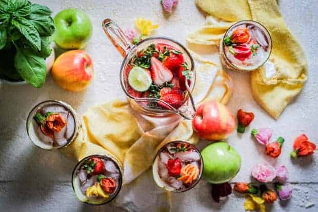 Top down view of a pitcher filled with sangria, 4 glasses are arranged around teh pitcher on teh table and are also filled with sangria. There is a white and yellow linen napkin on te table that wraps around the glasses. hibiscus blossoms and fresh pink and green apples are also arranged on the table. 