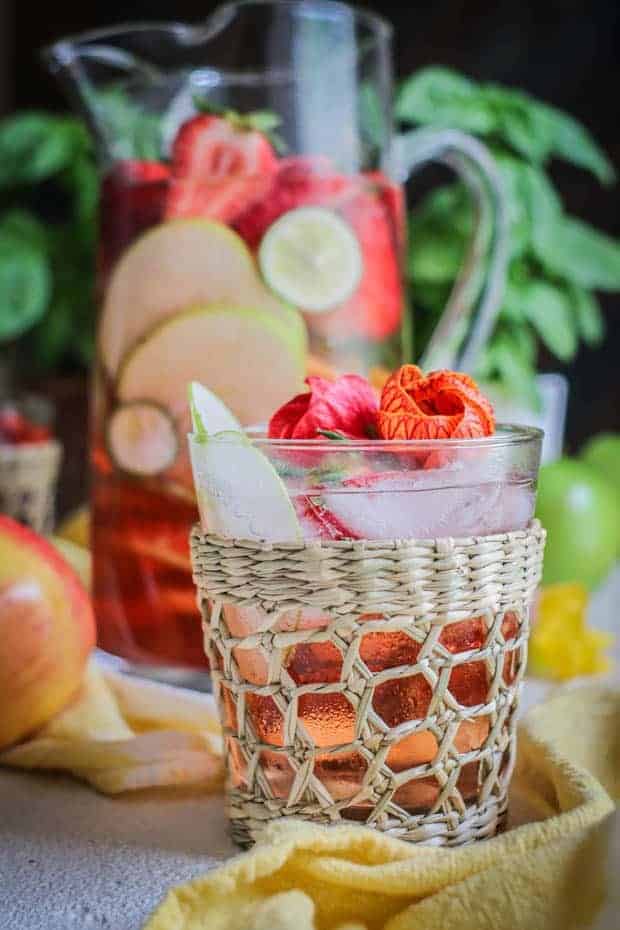 A clear cocktail glass with a woven ratan cover on it is filled with apple and strawberry slices, hibiscus blossoms, and apple and berry rosé sangria. Teh glass is in front of a large clear glass pitcher filled with the same fruits and beverage.
