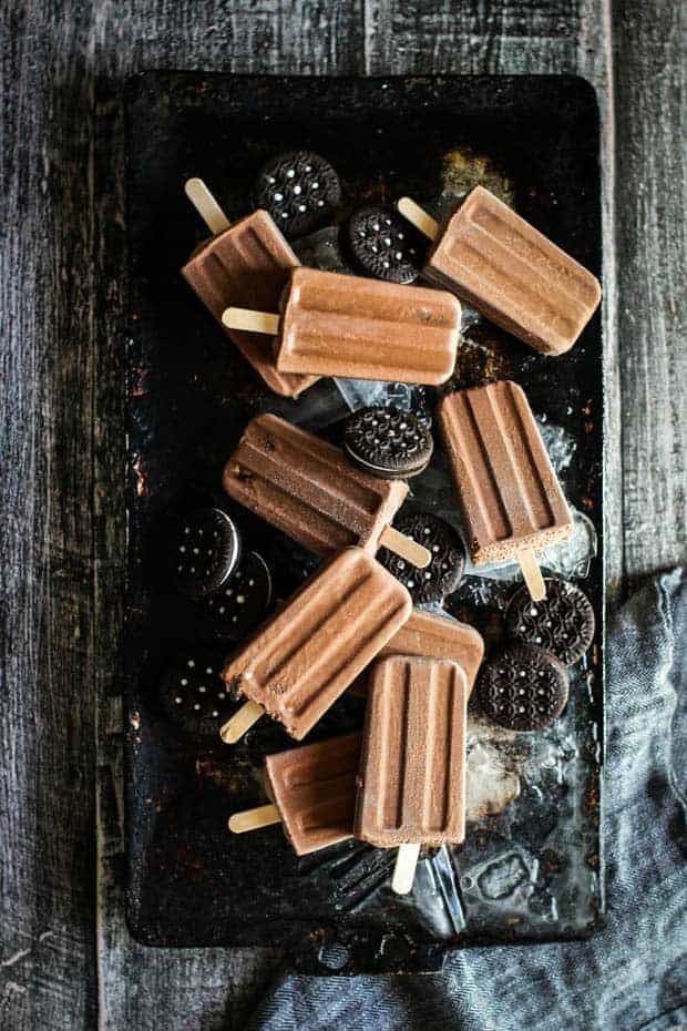 9 cookies and cream fudgesicles are scattered across a sheet pan that has a layer of frozen water and shattered ice on it. There are also chocolate sandwich cookies scattered amongst the fudgesicles