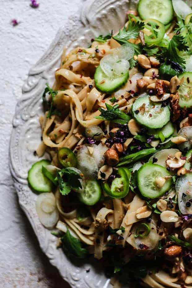 Side crop of a platter of No-Cook Peanut Butter Rice Noodle Salad on an off white back ground - teh =off-white pasta dish is topped with lots of green, crunchy vegetables.