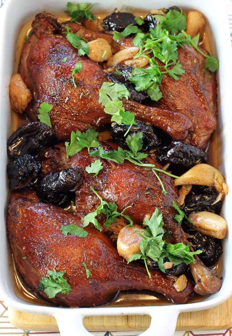 duck legs in a roasting pan with garlic cloves, dried plums, and cilantro.