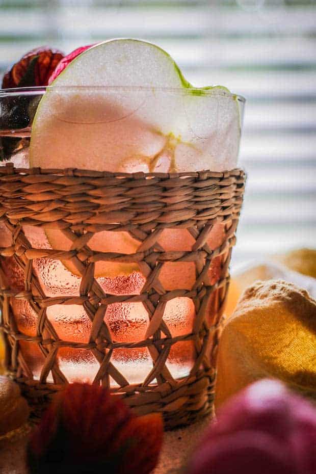 Up close photo of a glass of apple and berry rose sangria. The clear glass has a woven, ratan cup holder around it. You can see thin slices of apple, strawberry halves, hibiscus blossoms and ice cubes through the glass with the pink sangria.  There is a yellow linen napkin in the background. 