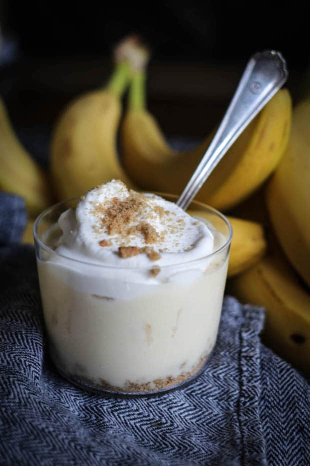 A bana cream pie pudding cup sits on a table with a silver spoon in it. There is a black and white houndstooth napkin underneath the cup and fresh bananas in the background. 