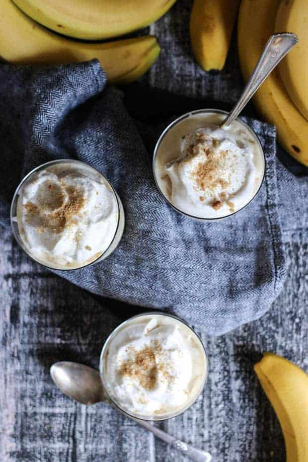 3 banana cream pie pudding cups sit on a table with fresh bananas and silver spoons.