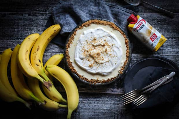 A banana cream pie with a ginger cookie crust is on a tael with fresh bananas next to it and a bottle of Premier Protein Bananas and Cream Protein Shake on the other side of the pie