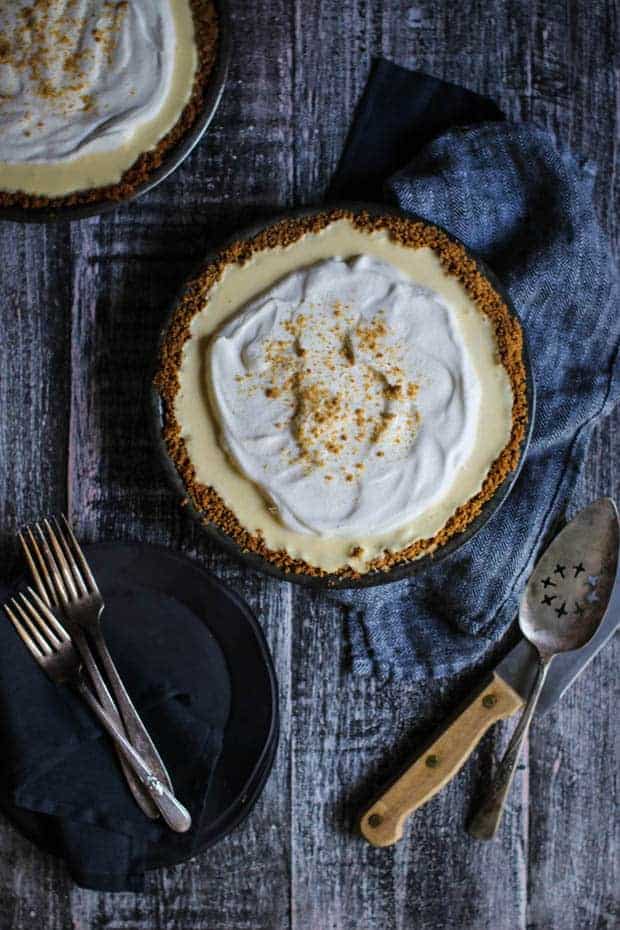 2 banana cream pies with ginger cookie crusts sit on a table next to a vintage knife with a wooden handle and a vintage pie server. There are also 2 small black plates stacked on the table next to the pie and they have forks on top of them.