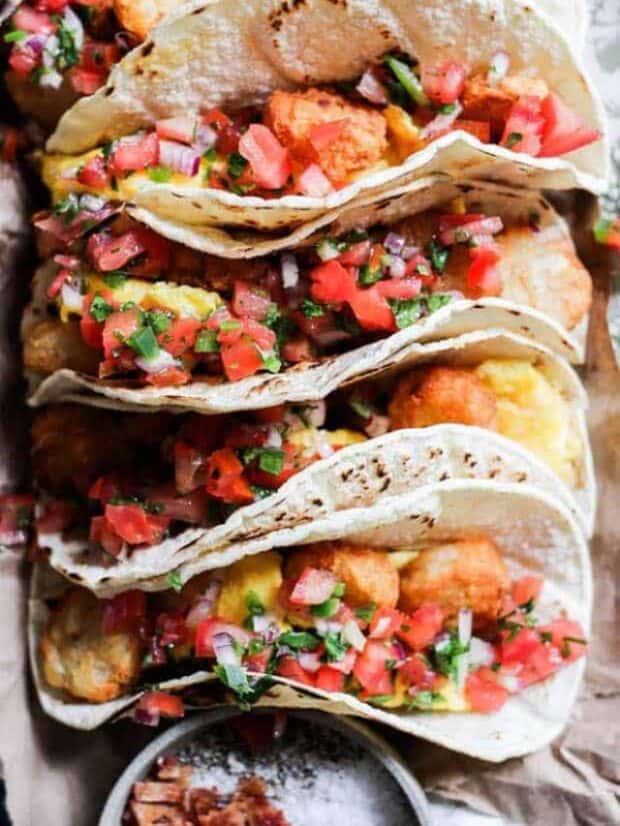 4 tacos, corn tortillas stuffed with scrambled eggs, tater tots, and pico de gallo. The tacos are arranged on a platter next to a bowl that has chopped bacon in it.