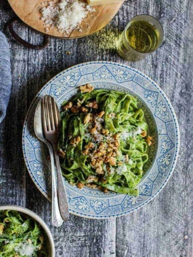 A large shallow bowl with a blue and cream decorative edge is piled high with noodles coated in a bright green pesto. There are walnuts and grated cheese on top and a fork and spoon on the side of the bowl. There is a glass if white wine next to the bowl.