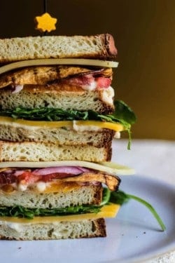 Close up image of 2 ultimate vegetarian club sandwiches staked on top of each other. It layers like this; br4ead, white bean hummus, cheddar cheese, arugula, bread, white bean hummus, yellow and red heirloom tomato slices, eggplant bacon slices, a slice of white cheddar cheese and another slice of bread.
