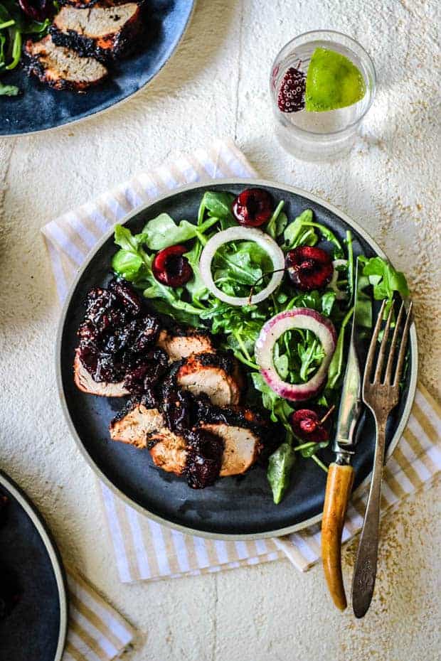 A black plate sits on a white table top over a pale cream and white striped cloth napkin. The plate has an aural salad with marinated red onion rings and fresh cherry halves. There are 3 slices of grilled pork tenderloin on teh plate topped with cherry ginger chutney.