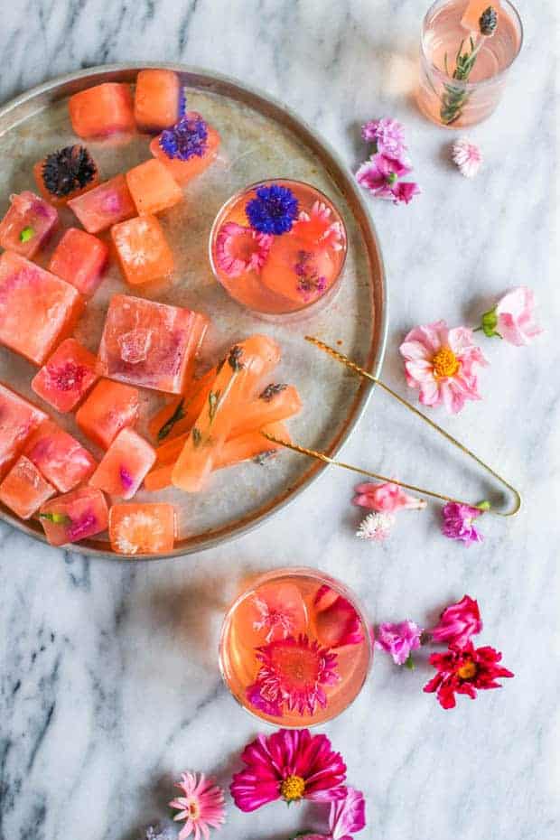 A round metal tray piled high with pink hued ice cubes that have colorful edible flowers frozen into them. There are also blooms of edible flowers scattered across the table. 
