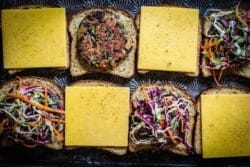 8 slices of bread on a baking sheet some are topped with slices of cheddar, some with veggie burger patties and some have the patties topped with a colorful slaw.