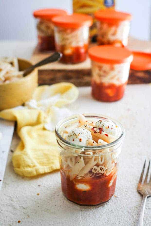 5 glass jars layered with pasta sauce, mozzarella balls, and penne pasta are on a table top. There is also a bowl of pasta on the table as well as a box of Pamelas gluten free pasta