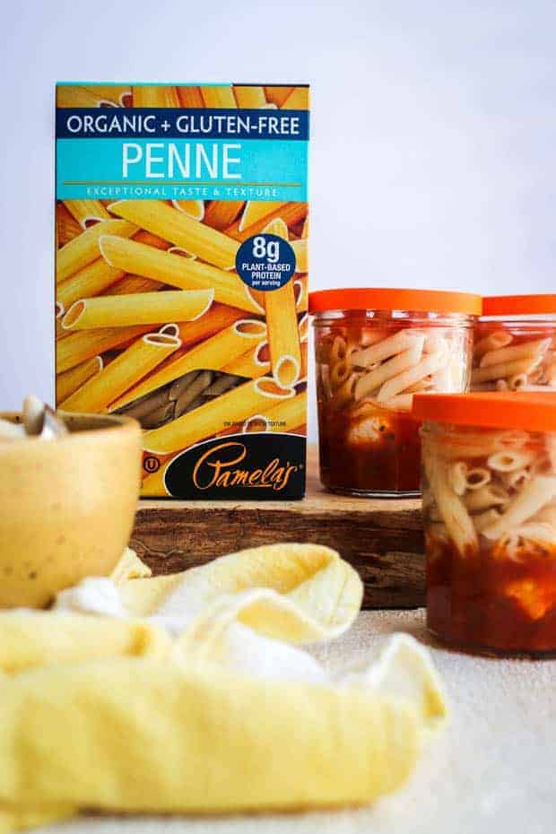 A box of Pamela's gluten free penne pasta on a wooden board next to small glass jars that are layered with marinara sauce, mozzarella balls, and gluten free pasta.