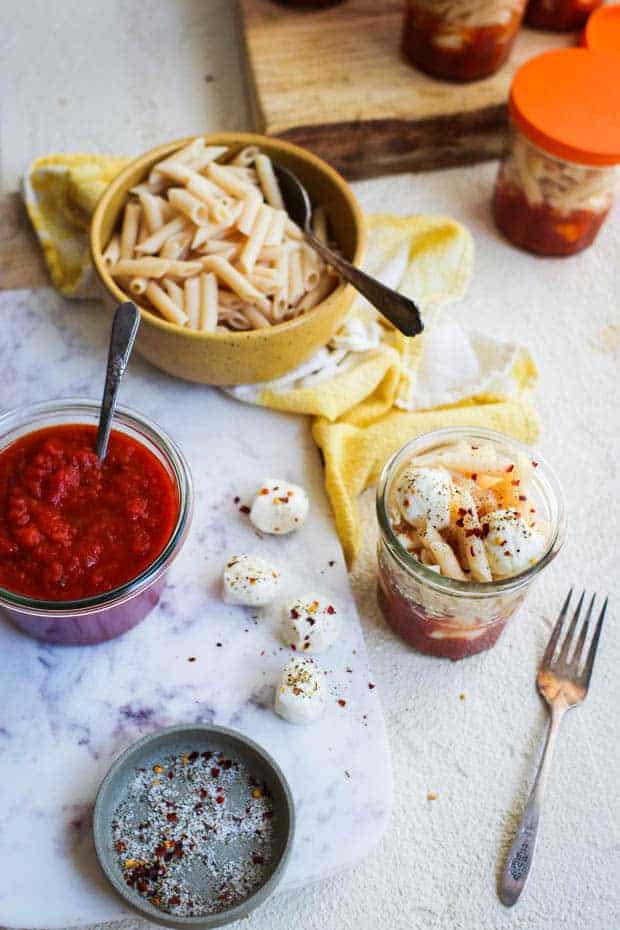 A prep scene where small glass jars are being filled with pasta sauce, mozzarella balls, and pasta. 