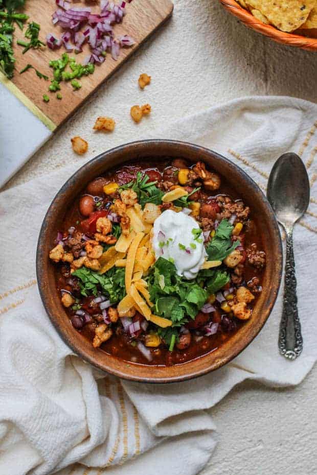 A bowl of hearty chili like soup is sitting on a cloth napkin. The soup is topped with shredded cheddar cheese, cilantro, diced red onions, homemade corn nuts, and sour cream. There is a basket of tortilla chips next to the soup. 