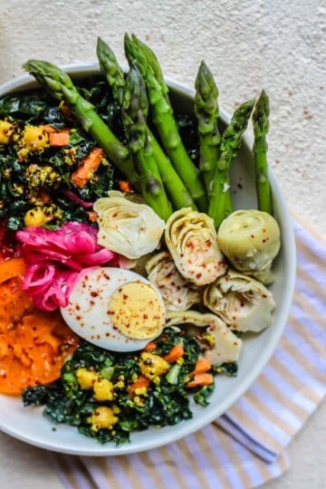 A vibrant bowl of kale salad with quinoa and chickpeas tossed in. There are heirloom tomato slices, pink pickled onions, deviled egg halves, blanched asparagus, and artichoke halves. The bowl is sitting on a white and tan striped napkin on a white background
