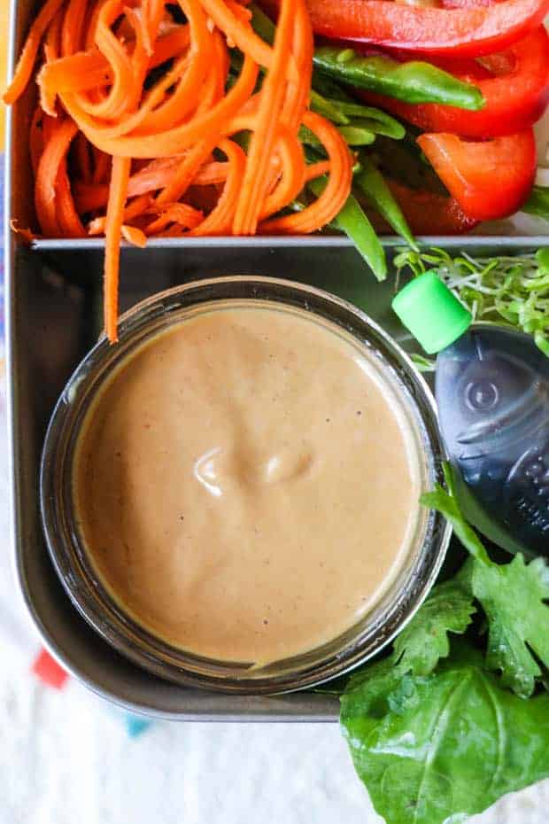 A jar of thai peanut dipping sauce in a bento box with shredded carrots and sliced red bell peppers and snow peas. there are a loud of herbs and a small container of soy sauce in the bento as well. 