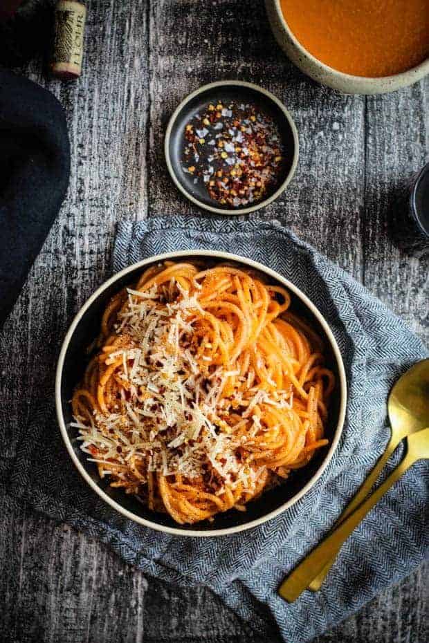 A bowl is piled high with pasta tossed in a red homemade marinara sauce. The pasta is topped with shredded Parmesan cheese and red pepper flakes. There is also a small bowl of sauce on the table and a glass of red wine.