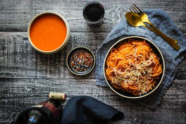 A dinner table scene with a bottle of wine and a bowl of pasta tossed in red sauce. There is a small bowl of red sauce on the table as well as a glass of red wine and silverware