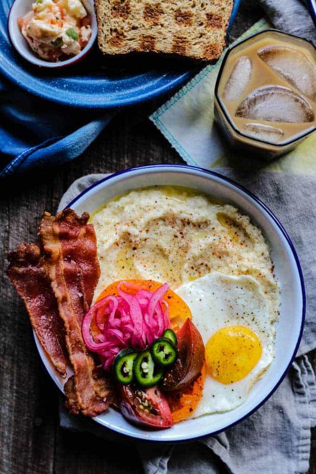 A white enamel bowl with a blue rim is filled with cheddar cheese grits then piled high with crispy bacon, sliced heirloom tomatoes, fresh jalapeño slices, pink pickled onions, and a perfectly fried sunny side up egg. 