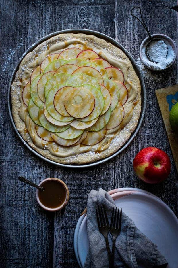 An entire gluten free Easy Apple Dessert Pizza is on a tbale next to plates and small bowls of caramel sauce and powdered sugar.