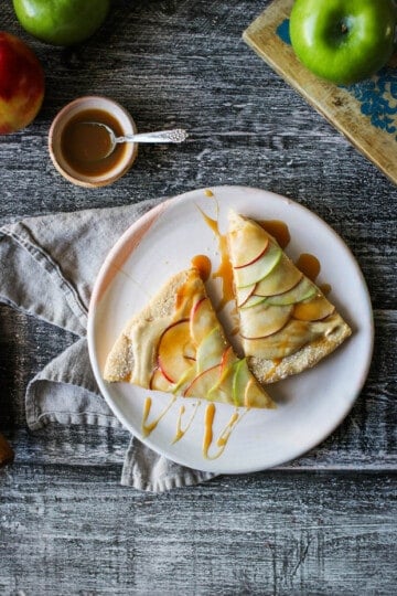 2 slices of easy apple desset pizza on a white plate - the slices have been drizzled with caramel. There are also green Granny Smith apples on the table.
