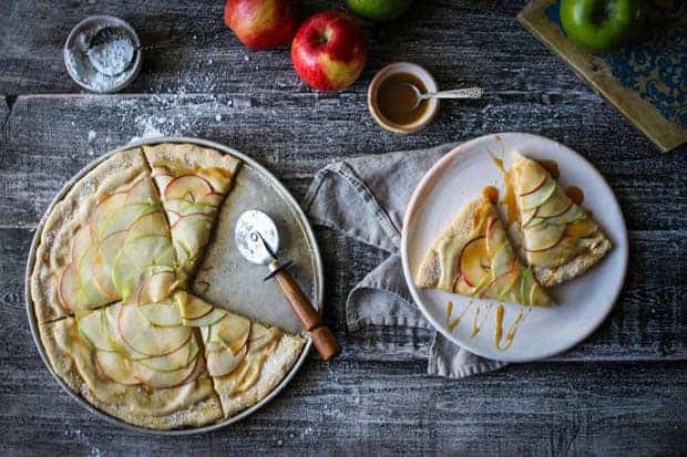 A table top with an Apple Dessert Pizza cut into slices. There are 2 slices pulled on to a plate from the pan and they are drizzled with caramel. There are also fresh red and green apples on the table.