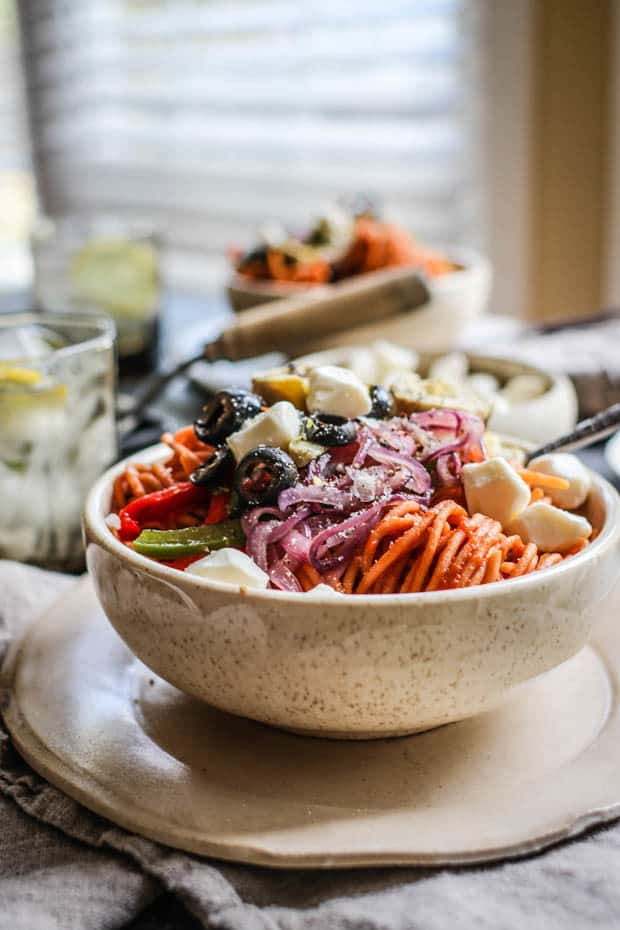 A bowl of red lentil pasta that has been tossed with pizza sauce and then piled high with roasted peppers, onions, artichokes, and black olives.