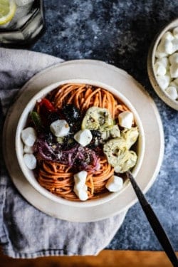 A top down photograph of a bowl of red lentil pasta tossed with pizza sauce and topped with roasted bell peppers, onions, artichokes, black olives, and mozzarella balls