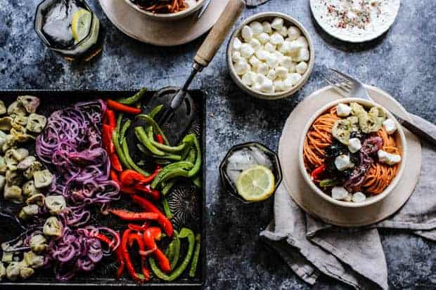 A sheet pan of roasted peppers and onions next to a bowl of red lentil pasta tossed with pizza sauce then topped with roasted vegetables like bell peppers, roasted onions, artichokes, and black olives. Topped with mozzarella balls.