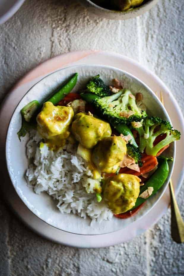 A bowl filled with jasmine rice then topped with stir fries broccoli, cauliflower, snow peas, and carrots. On top of the vegetables are 4 coconut curry meatballs smothered in yellow curry sauce