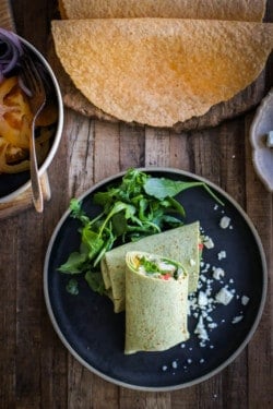 A brown butter chicken wrap on a black plate next to a platter of toufayan gluten free wraps. The wrap itself is spinach flavored so it is green and vibrant.