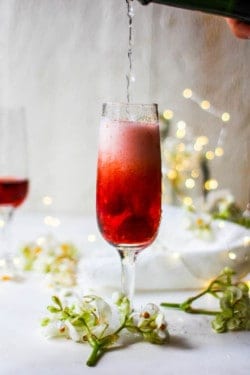 a wine glass is being filled with fizzy white wine. The glass has red aspberries and a red hied liquid in it already