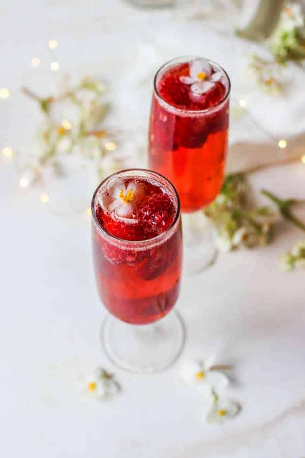 2 champagne glasses filled with a beautiful red drink, there are raspberries floating in the glass and small white flowers an dligts on the table around it