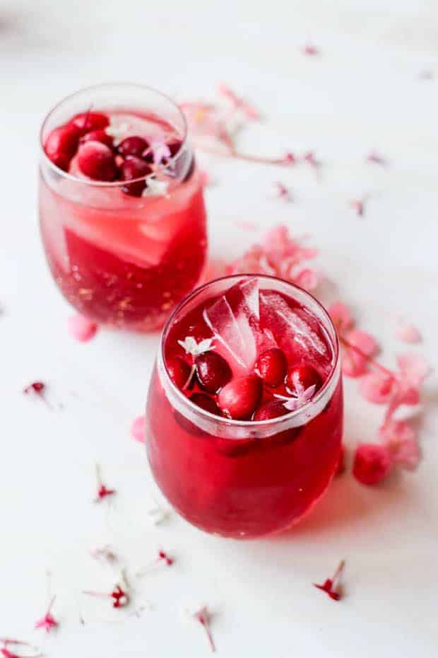 2 stemless wine glasses are o a white table filled with a red cocktail and garnished with frozen cranberries and pink edible flowers