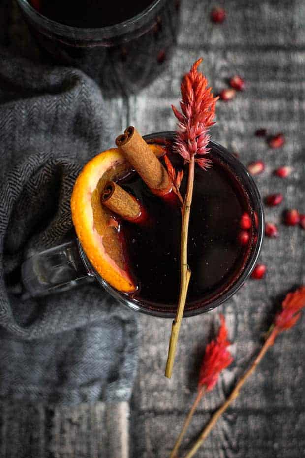 Lookin down into a mug of warm mulled red wine. There is a cinnamon stick, orange slice, and pomegranates floating in the mulled wine. 