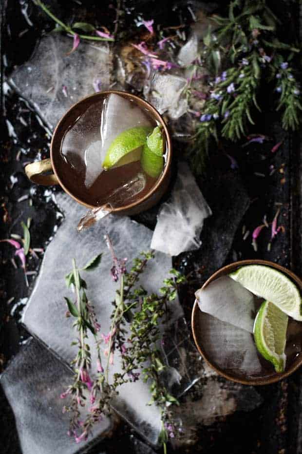 top down image looking into 2 copper mugs filled with a cocktail and lime wedges. there are fresh flowers scattered around the table and sheets of ie underneath the mugs.