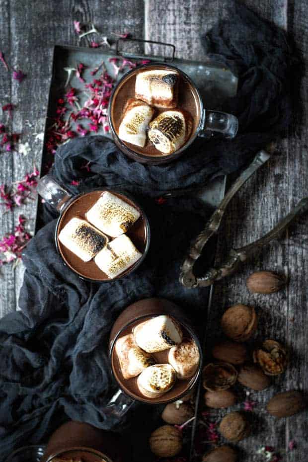 3 mugs of red wine hot chocolate are in a metal tray. Each mug is topped with toasted marshmallows. There is a dark colored cloth on the tray with the mugs of hot chocolate and there are walnuts in the shell, a vintage nut cracker an dpink flowers scattered all over the table.