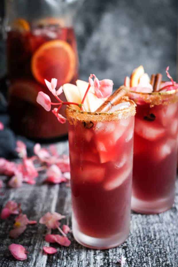 2 glasses of red apple sangria are styled on a table top - they are loaded wit hfresh apple slices, cinnamon sticks, lotc of ice, pink edible flowers, and a delectable brown sugar rim there is a pitcher of sangria in the background 